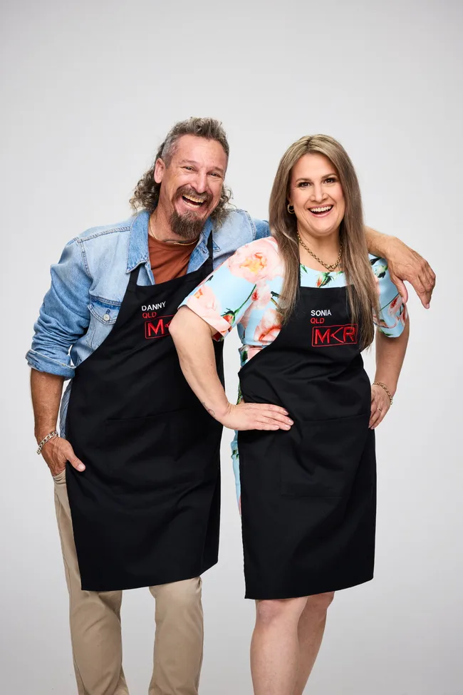 Two people wearing My Kitchen Rules aprons, smiling and posing with their arms around each other.