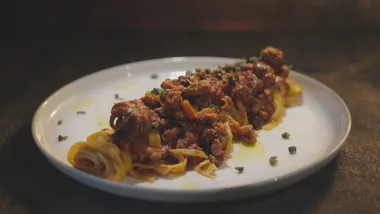A plate of pasta with rich tomato meat sauce garnished with herbs, set on a dark surface.