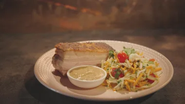 A plate with crispy pork belly, salad with cherry tomatoes, and a small bowl of sauce.