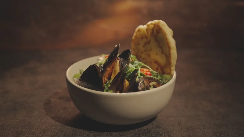 Bowl of mussels served with greens and a slice of bread on a dark background.