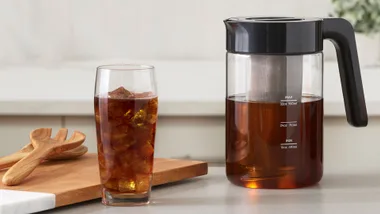 A glass of iced tea with a pitcher on a kitchen counter, wooden utensils on the left.