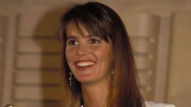 Smiling woman with long brown hair and earrings, wearing a white top.
