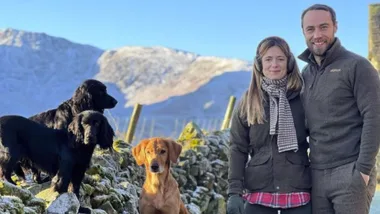 Couple smiling with three dogs on a snowy mountain pathway under a clear blue sky.