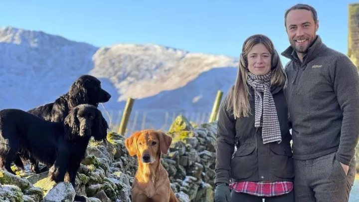 Couple smiling with three dogs on a snowy mountain pathway under a clear blue sky.