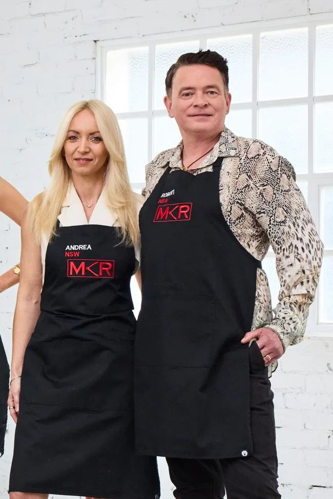 Two people wearing aprons with "MKR" logo stand in front of a window in a bright room.