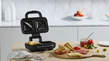 Open sandwich maker with triangular slots, sandwiches on cutting board, ingredients like salami and cheese nearby.