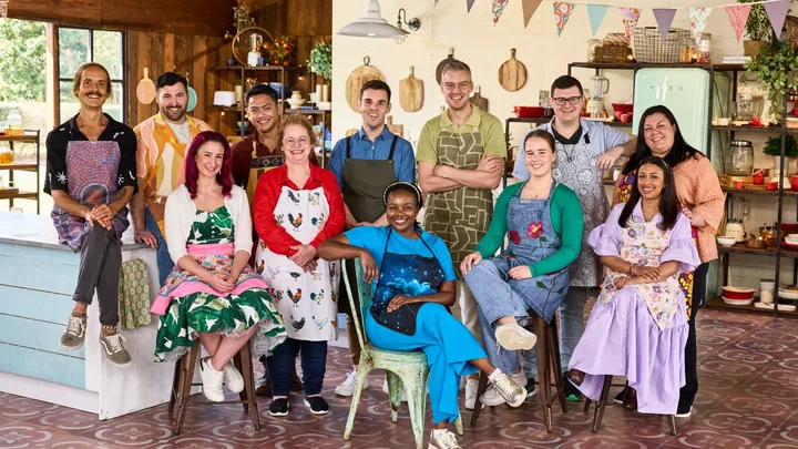 A diverse group of contestants from The Great Australian Bake Off 2023, wearing colorful aprons, smiles in a cozy kitchen setting.