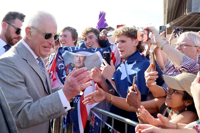 Man in gray suit and sunglasses greeting excited crowd holding flags and phones, with a prominent building in the background.
