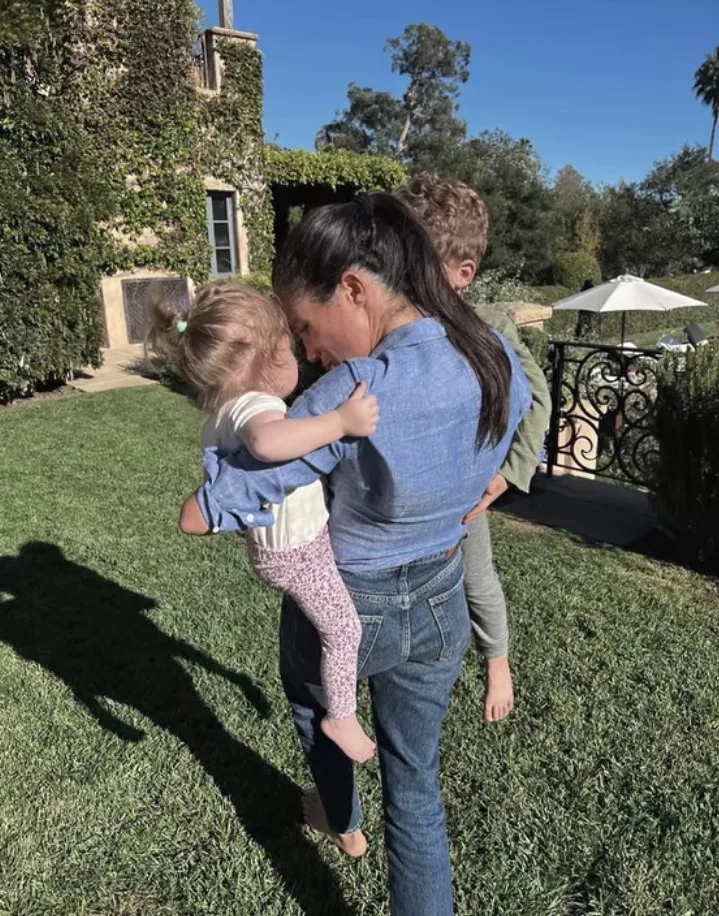 Meghan carrying her children Archie and Lilibet whose faces are turned away from camera.