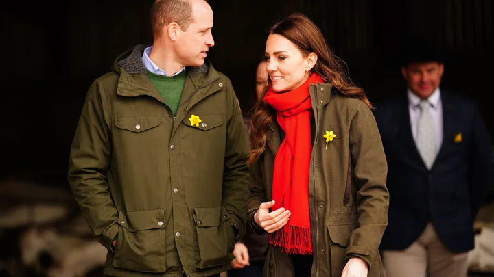 A man and a woman wearing matching green jackets with daffodil pins, the woman also wearing a red scarf, walking together.