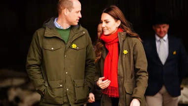 A man and a woman wearing matching green jackets with daffodil pins, the woman also wearing a red scarf, walking together.
