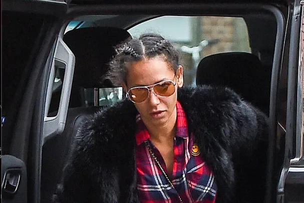 A person in sunglasses and a plaid shirt exits a vehicle, wearing a black coat with a fur collar.