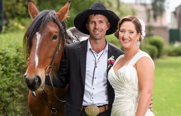 A couple in wedding attire with a horse, standing outside on a sunny day, both smiling at the camera.
