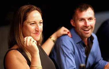 Two people smiling and seated at a table during a social event, one with a hand on the other's shoulder.