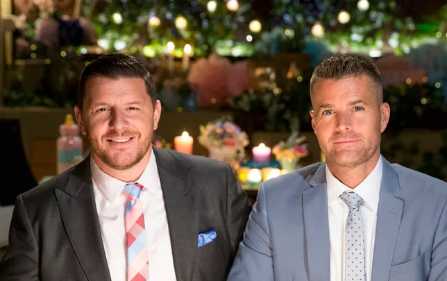 Two men in suits sitting at a table with decorative candles and flowers in the background.