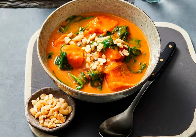 Bowl of peanut stew with sweet potato and spinach, topped with chopped peanuts, next to a bowl of peanuts.
