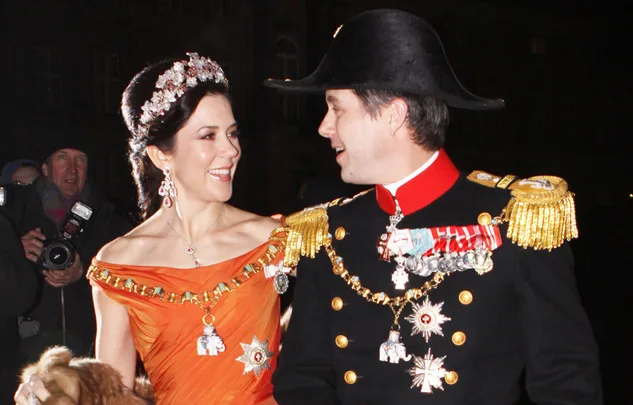 A man in military attire and a woman in an orange gown with a tiara smile at each other at a formal event.