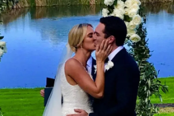 A bride and groom share a kiss in front of a scenic lake and floral arch during their outdoor wedding ceremony.