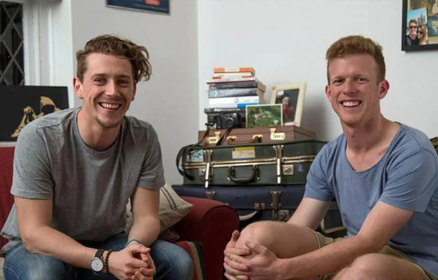 Two smiling men seated in a cozy room with books and a camera on a table behind them.