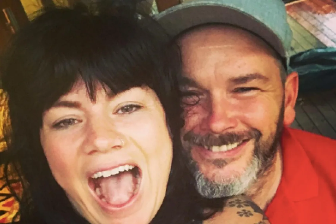 A smiling woman with dark hair and a man in a cap, close together for a selfie.