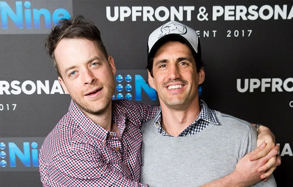 Two men smiling and embracing at an event in front of a backdrop with "Nine" and "Upfront and Personal" logos.