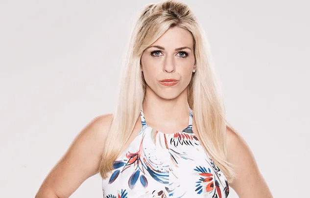 A woman with long blonde hair in a floral top poses against a light gray background.