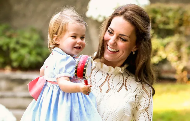 Mother smiling, holds young girl in blue dress with toy, outdoor setting.