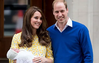 A couple smiling outside a building, with the woman holding a newborn wrapped in a white blanket.