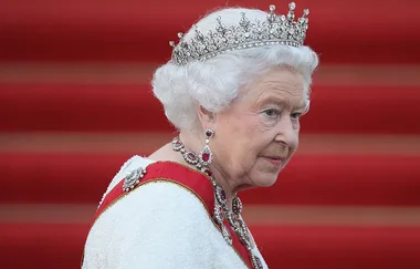 A person in royal attire and a jeweled crown against a red carpet backdrop.