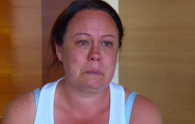 A woman with teary eyes looks emotional, sitting indoors against a wooden background, wearing a white tank top.