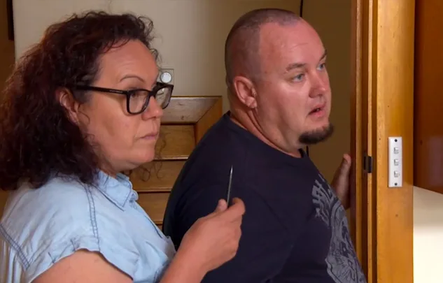 Two people in a room, one holding an object, both looking intently in the same direction.