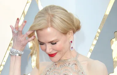 Blonde woman in an elegant gown with red lipstick and jewelry, adjusting her hair, against a glamorous backdrop.