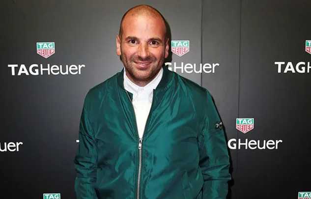 A person in a green jacket poses in front of a TAG Heuer backdrop.
