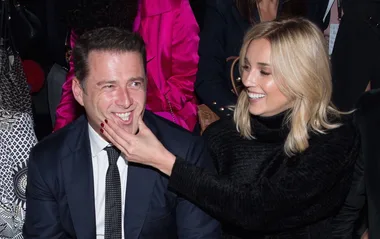 A man in a suit and a woman in a black top smiling, seated in the front row at a fashion event.