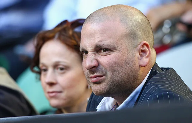Man in a suit making a grimacing expression, seated beside a woman with a neutral expression.