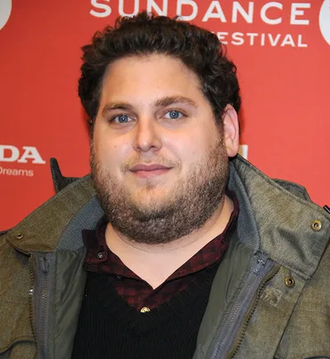 Actor at Sundance Festival wearing a jacket and checkered shirt, smiling, red background.
