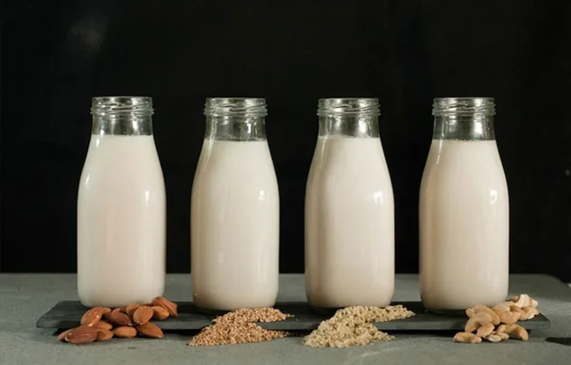 Four glass bottles of nut and seed milk with almonds, quinoa, sesame seeds, and cashews on a table.