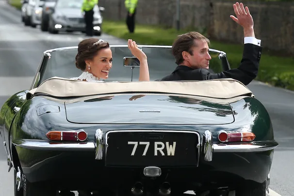 Bride and groom waving from a vintage convertible car, license plate "77 RW", with police escort in the background.