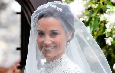 Bride smiling under a veil on her wedding day, wearing a tiara and lace dress, with a floral background.