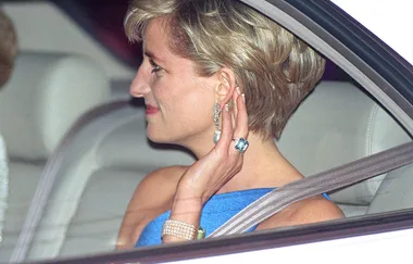 A woman in a blue dress, sitting in a car, wearing elegant jewelry and an aquamarine ring.