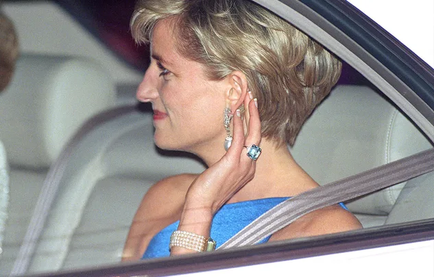 A woman in a blue dress, sitting in a car, wearing elegant jewelry and an aquamarine ring.