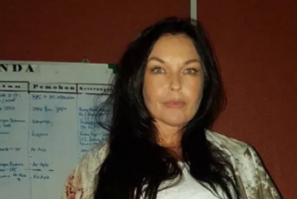A woman with long dark hair stands in front of a whiteboard with notes.