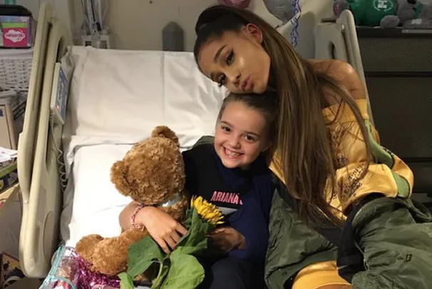 Ariana Grande hugs a young girl in a hospital bed, both smiling with a teddy bear and a sunflower.