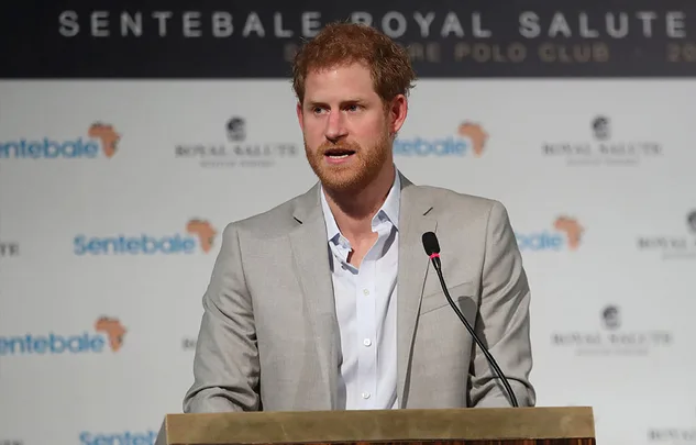 Prince Harry speaking at Sentebale Royal Salute Polo Cup event, podium and backdrop visible.