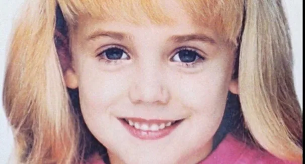 A young girl smiling brightly with long blonde hair, wearing a pink top.