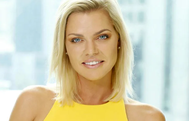 Blonde woman in a yellow top smiling, with a blurred cityscape in the background.