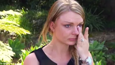 A woman with blonde hair wipes her tears in a garden setting, looking emotional.