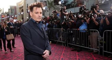 A person in a suit poses on a red carpet, with a crowd of photographers taking pictures in the background.