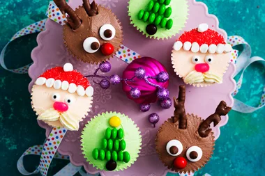 Christmas cupcakes decorated as Santa, reindeer, and trees on a purple plate with a pink bauble.
