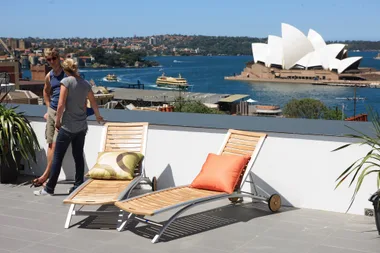 Two people talking on a rooftop terrace overlooking Sydney Opera House with deck chairs in the foreground.